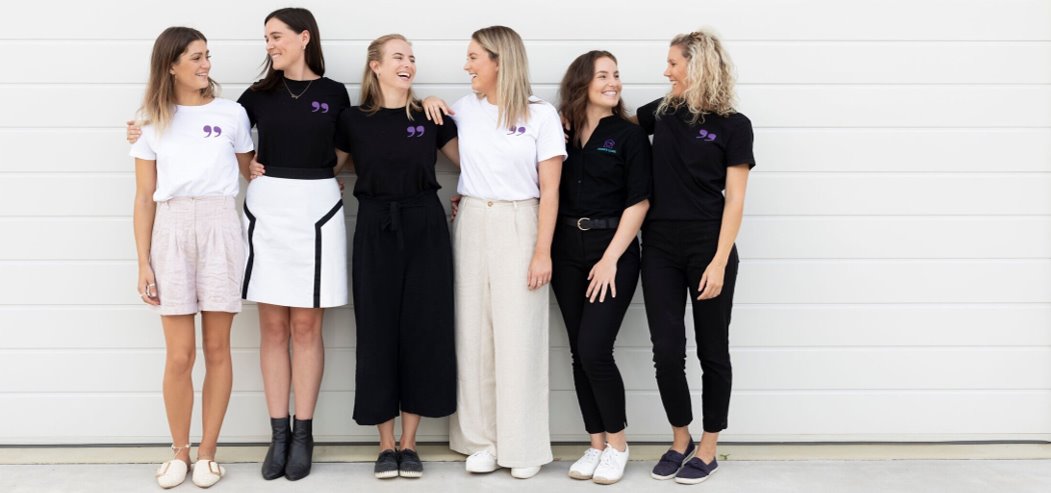 A group of 6 women, hands over each other’s shoulders and smiling at one another.