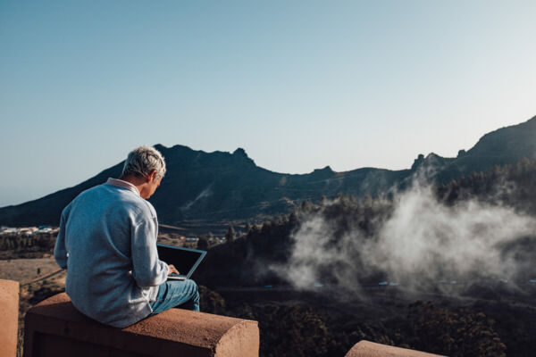 Photo shows a man with light blonde hair, blue shirt using his laptop with view of mountains in the distance.