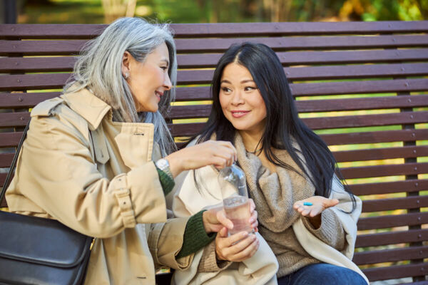 Photo shows a cheerful Asian woman with grey hair helping a younger Asian woman with long dark hair to take a pill.